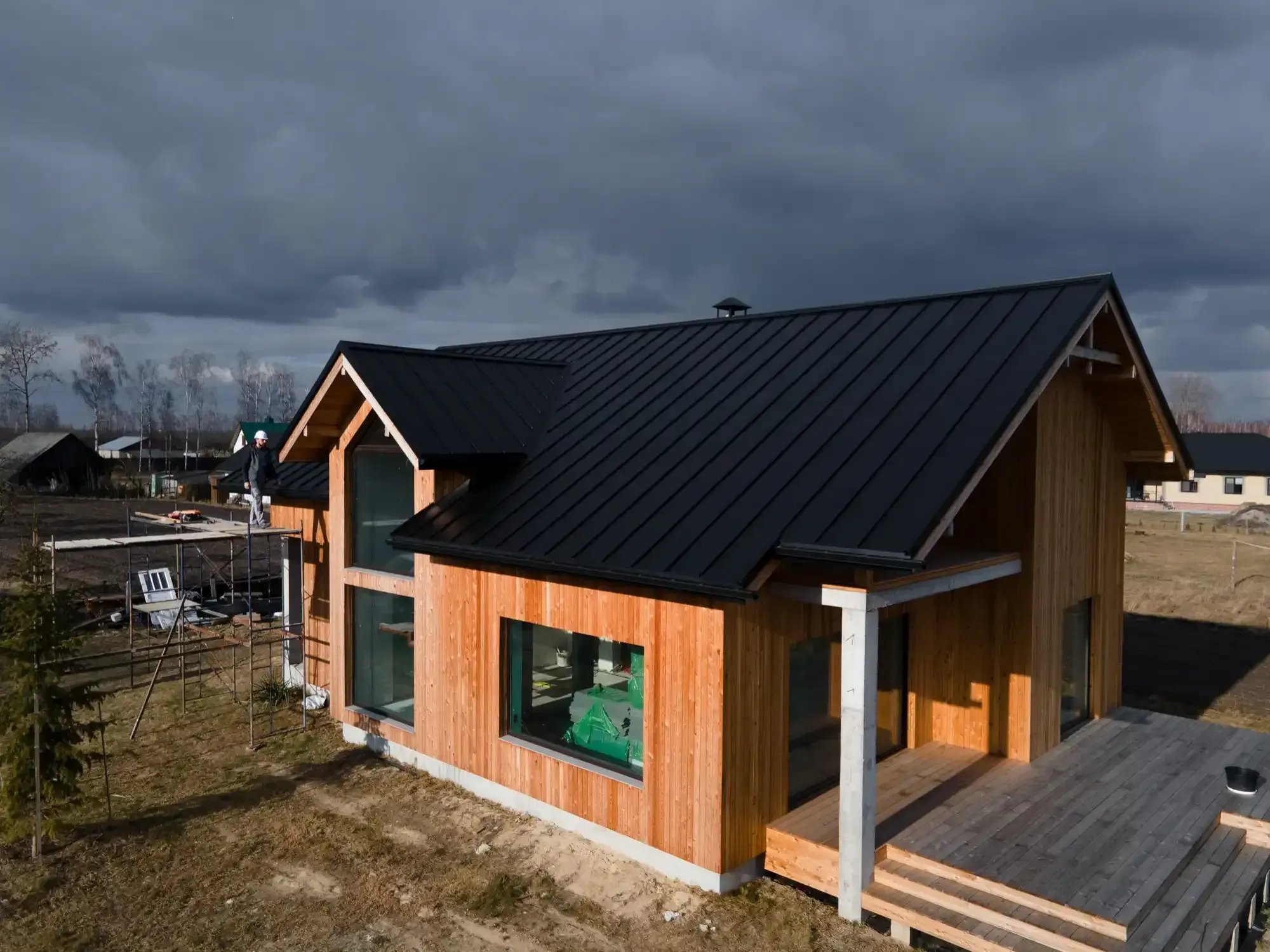 Wooden house with black roof, large windows, and porch under a cloudy sky.
