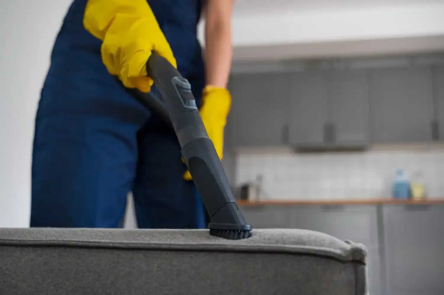Person in yellow gloves vacuuming a gray sofa in a kitchen.