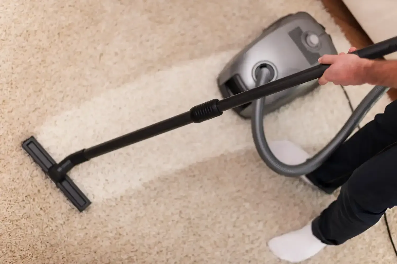Person vacuuming a beige carpet with a gray vacuum cleaner; creating a clean path.