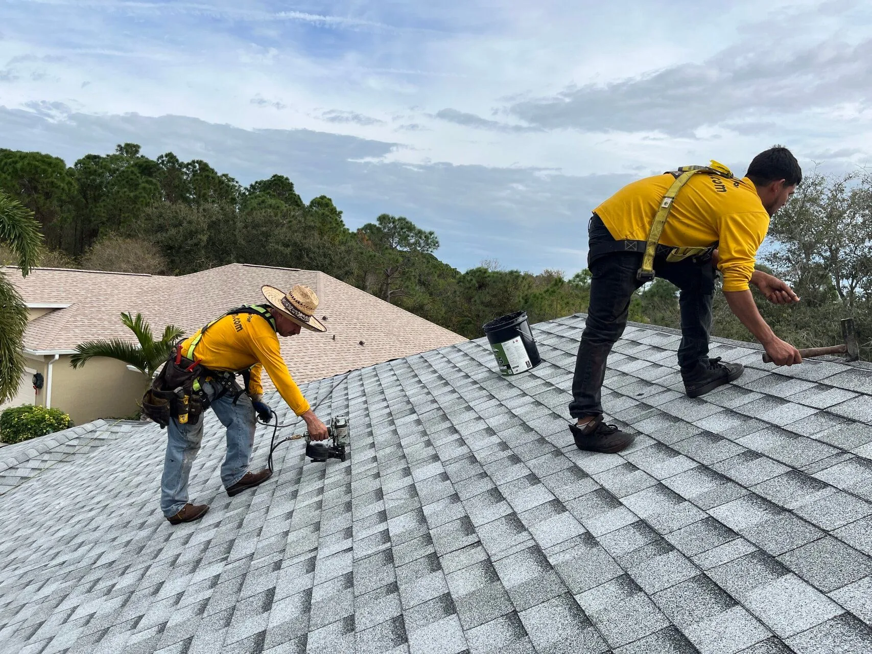 Two roofers installing gray shingles on a residential roof under a cloudy sky.